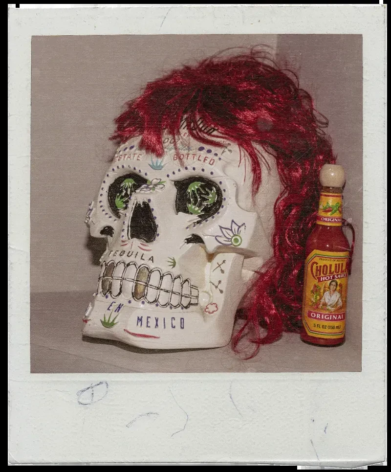Instant photo shows decorative red-haired skull beside Cholula hot sauce bottle.