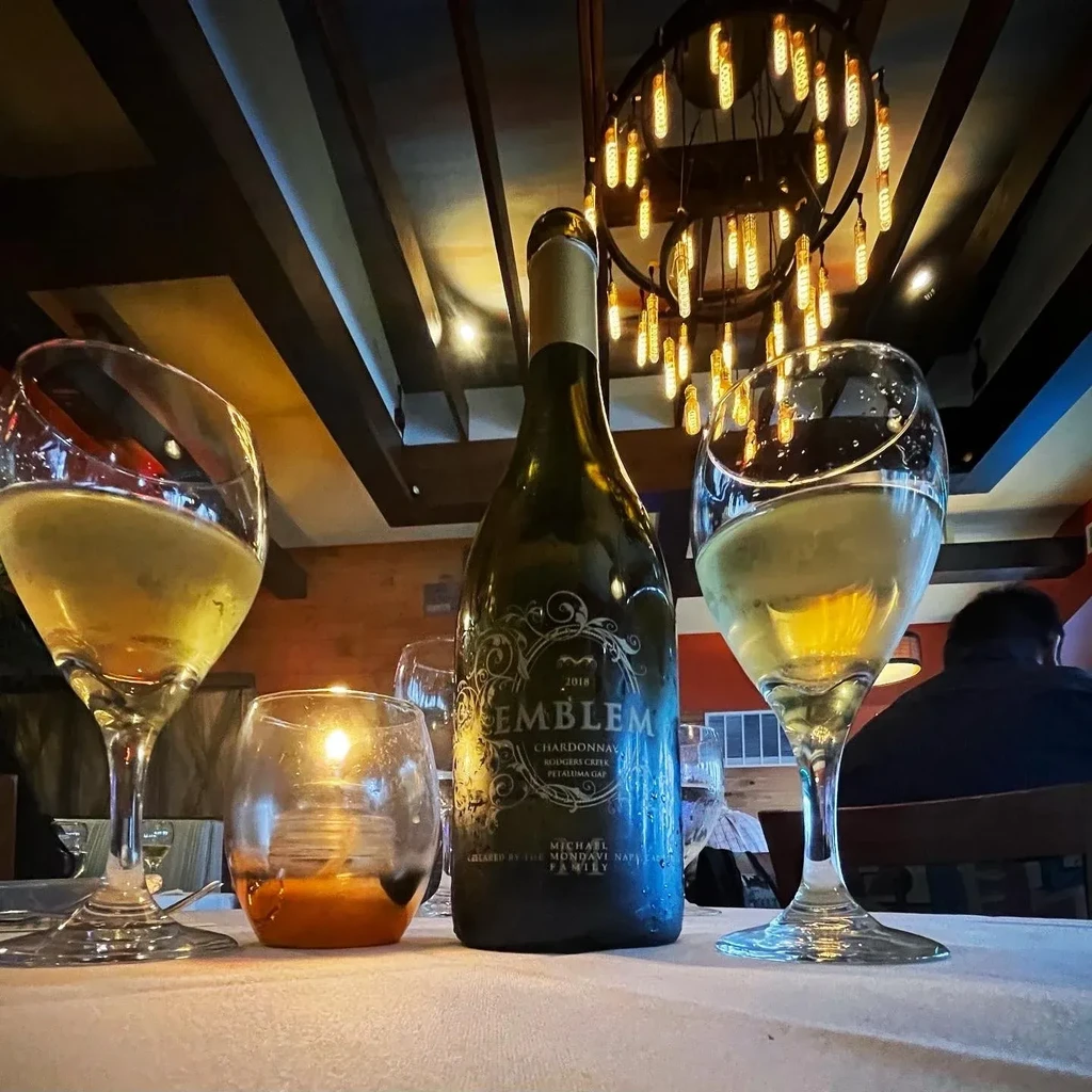 Emblem Chardonnay bottle, two glasses, and candle on restaurant table.
