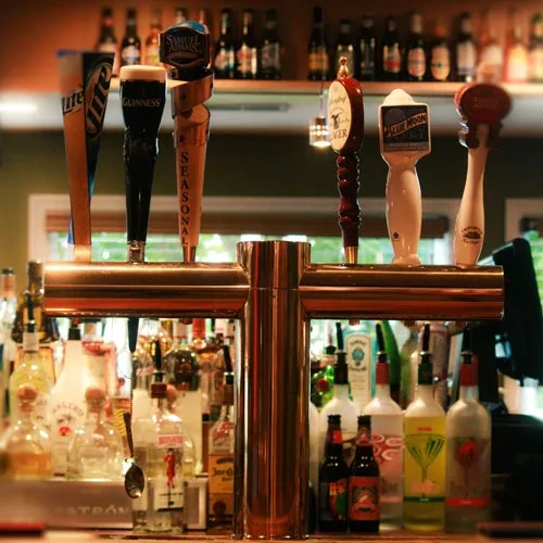 Five brass beer taps with brand names and liquor bottles in a bar.