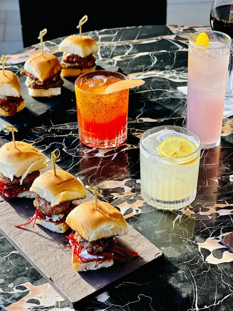 Sliders with skewers and three vibrant cocktails on a dark marble table.