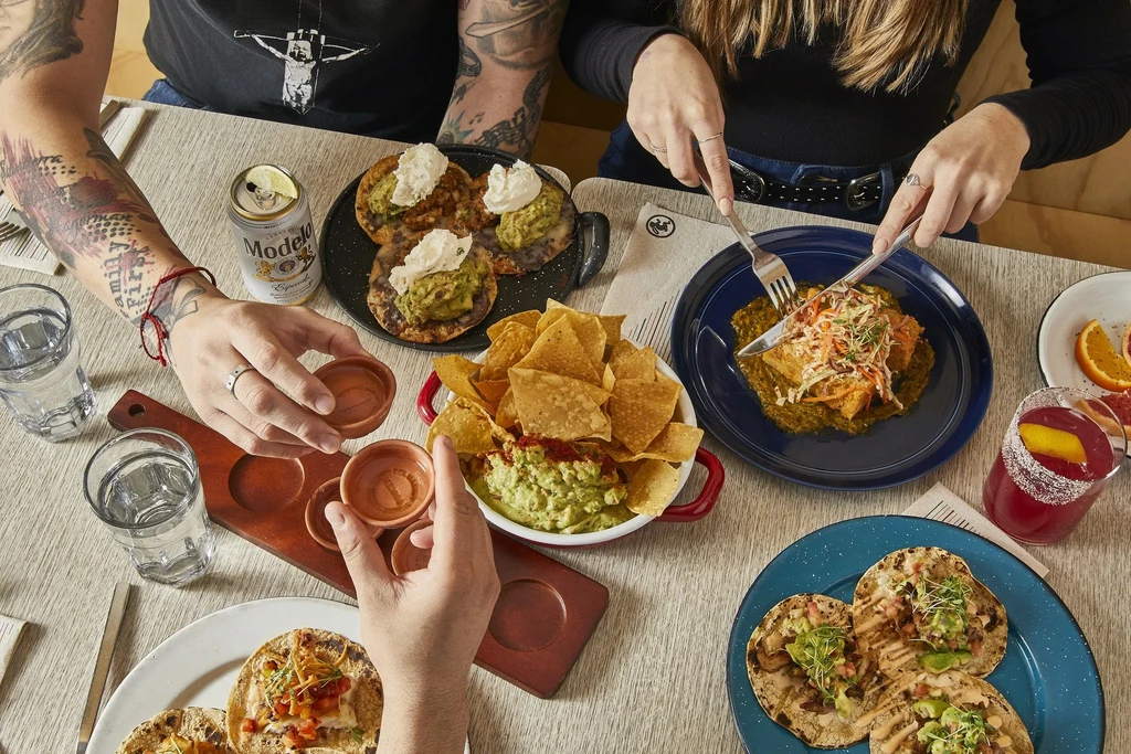 Overhead shot of a restaurant table with tacos, guacamole, drinks, and hands.