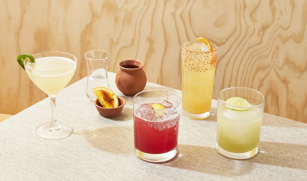 Five garnished beverages, a pitcher, and a pot on a light table.