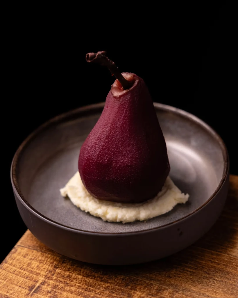 Deep red poached pear with stem on creamy base in bowl.
