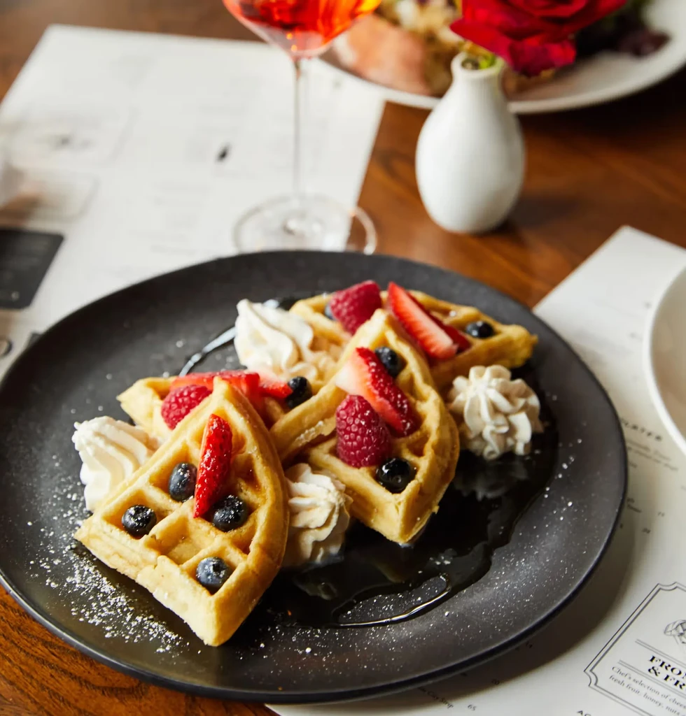 Golden-brown waffles with fresh berries, whipped cream, syrup, and powdered sugar.