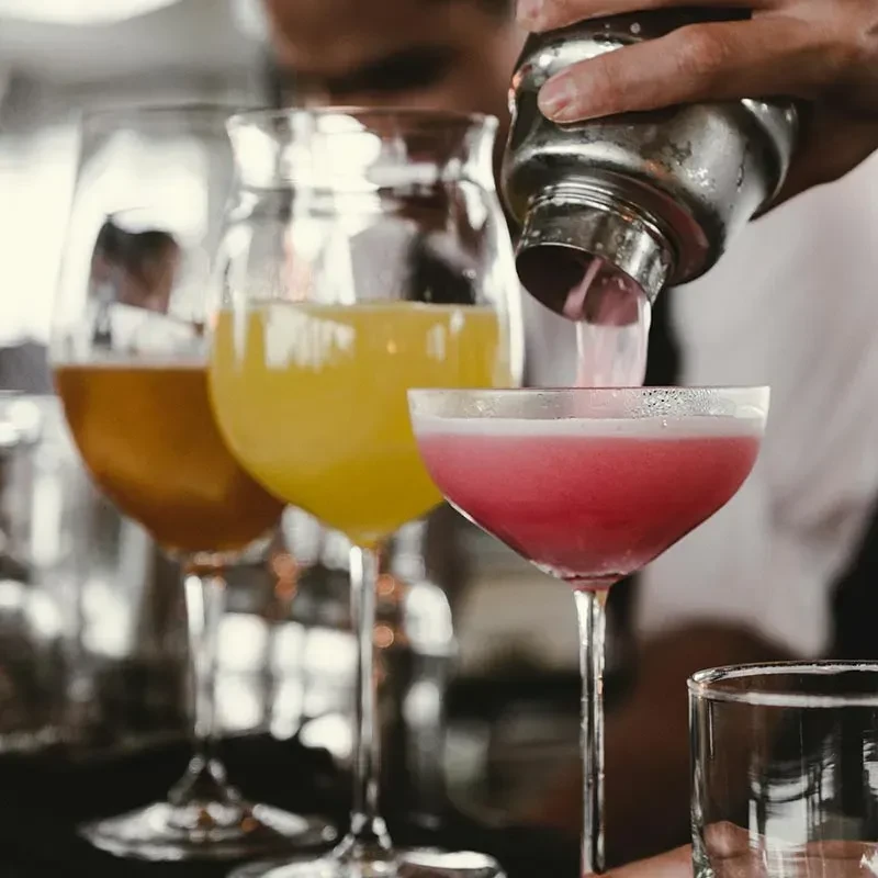 Hands pour bright pink cocktail from shaker into coupe glass.