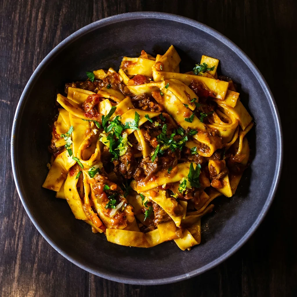 Close-up of wide pasta with meat ragu and herbs in a dark bowl.