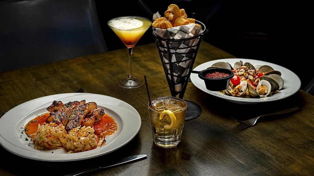 Dark wooden table with grilled meat, cocktails, fried calamari, and clams.