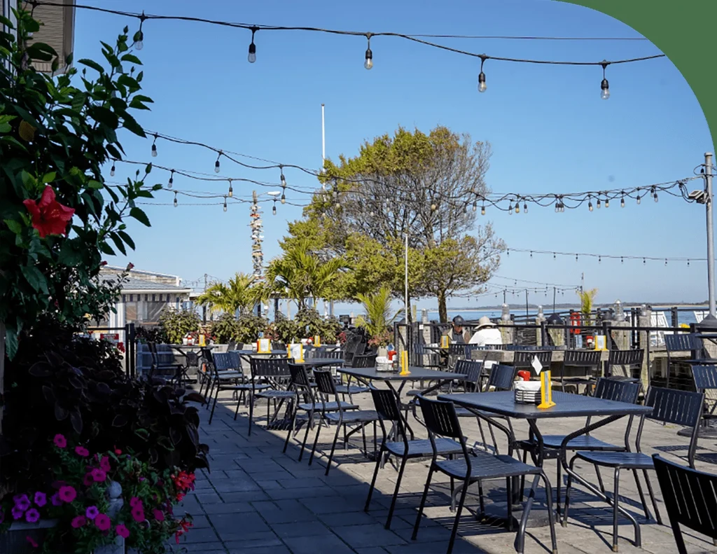 Outdoor waterfront restaurant patio with tables, string lights, and colorful plants.