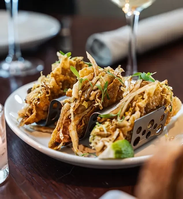 Three crispy tacos in metal stands, filled with vegetables and lime.