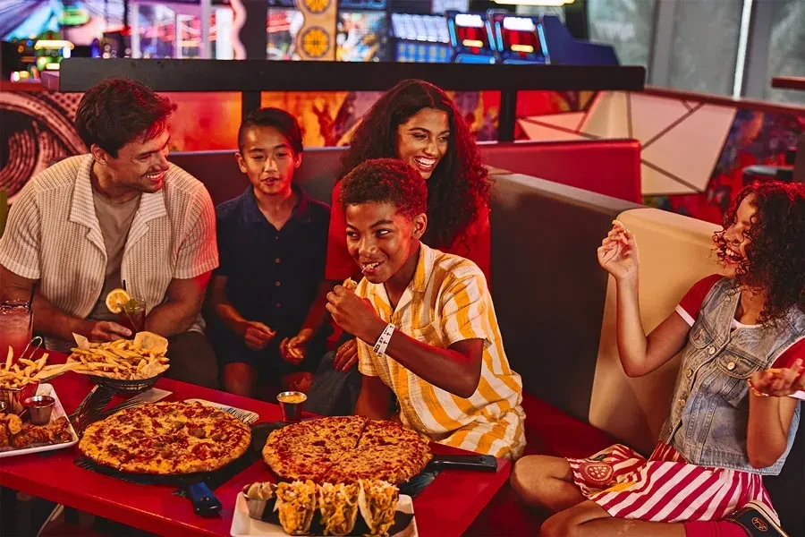 Family of five laughing, sharing pizzas and food at a restaurant table.