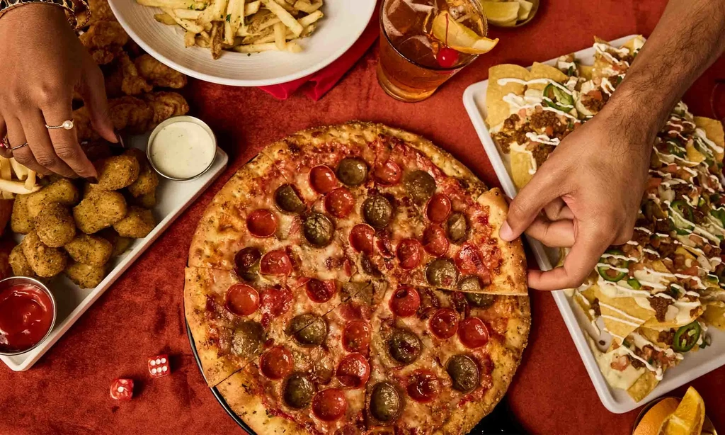 Hands share pizza, nachos, and fried foods on a deep red table.