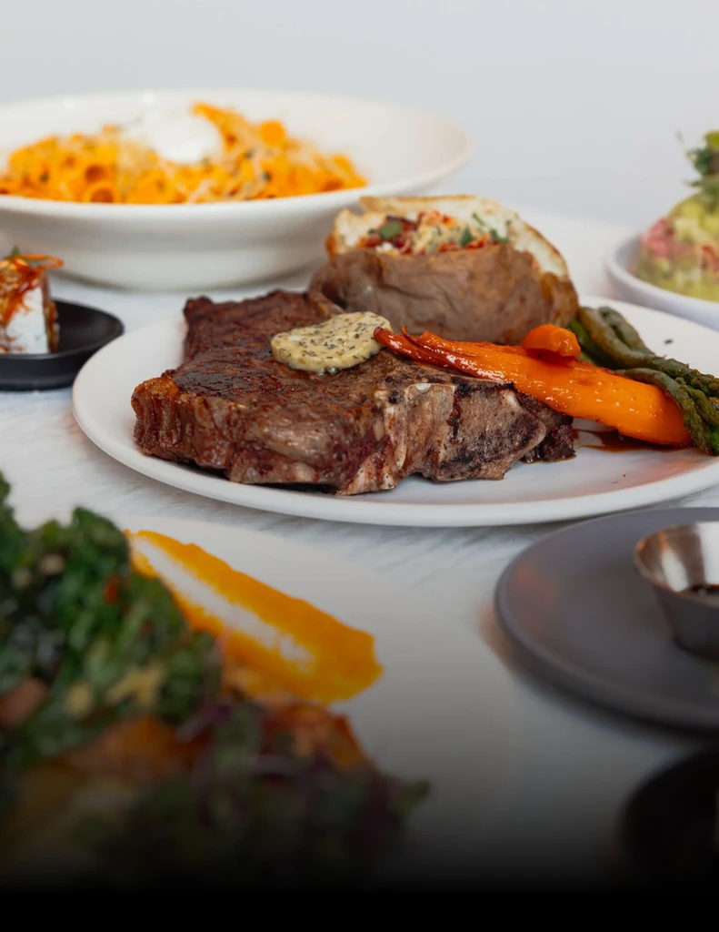 Restaurant table with steak, vegetables, loaded potato, pasta, and other dishes.