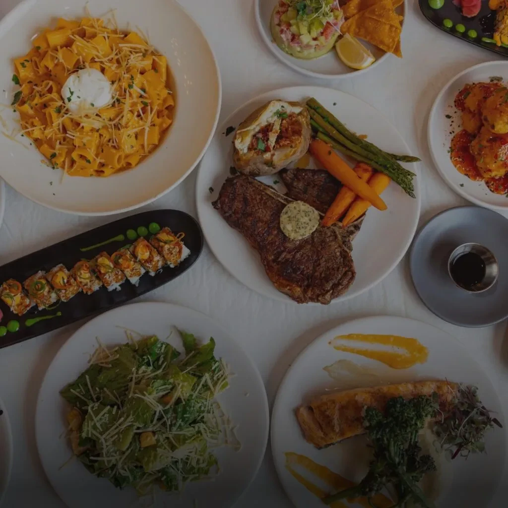 Table laden with diverse plates of pasta, steak, and sushi.