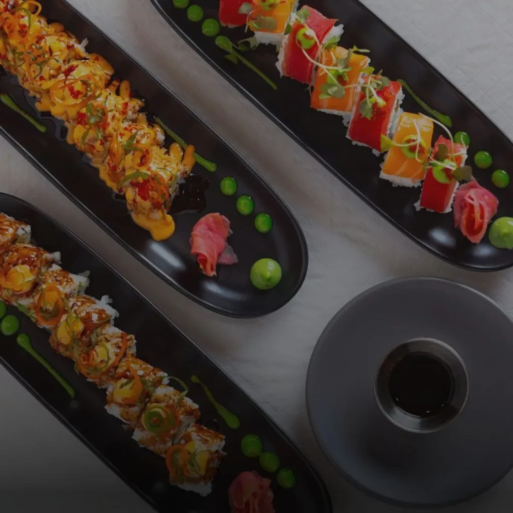Overhead, three black plates hold elaborately garnished sushi rolls and soy sauce.