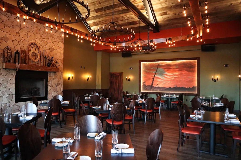 Rustic restaurant interior with stone fireplace, wooden ceiling, and set tables.
