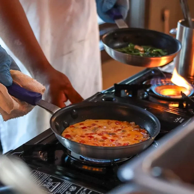 Two gloved hands cook colorful omelette and green vegetables in pans.