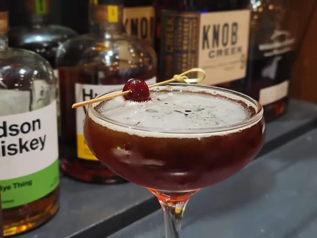Dark cocktail in a coupe glass with frothy head and red cherry.