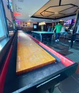 Shuffleboard table extends through a dimly lit bar with pool and people.
