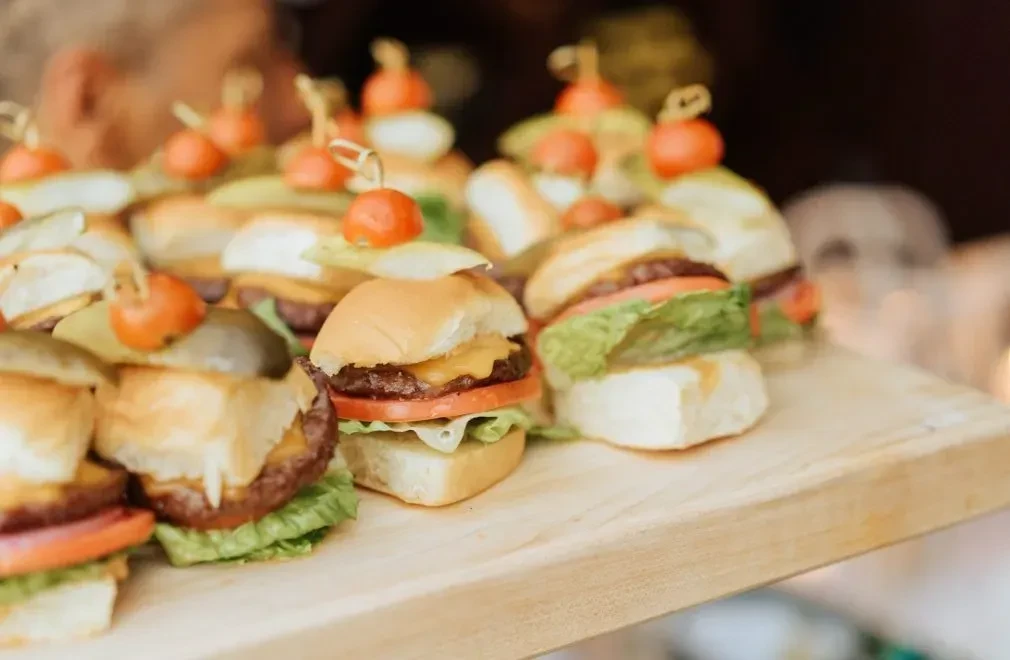 Mini beef sliders with cheese, tomato, lettuce, and pickle on a board.
