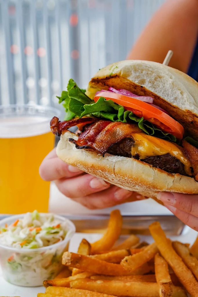 Hands hold a bacon cheeseburger, fries, and coleslaw.