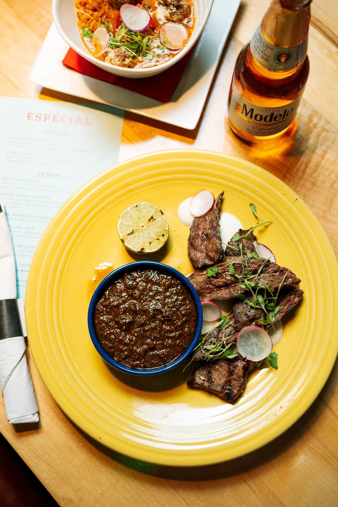 Grilled steak, meatball stew, and Modelo beer on a wooden table.