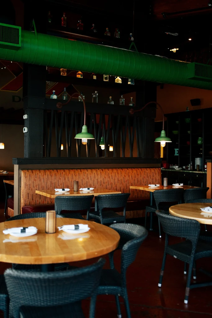 Dim restaurant with wooden tables, orange booth, bar, green duct.
