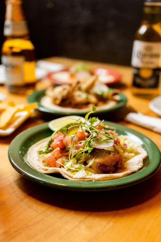 Detailed view of a fried taco with salsa, microgreens, lime, and beer.
