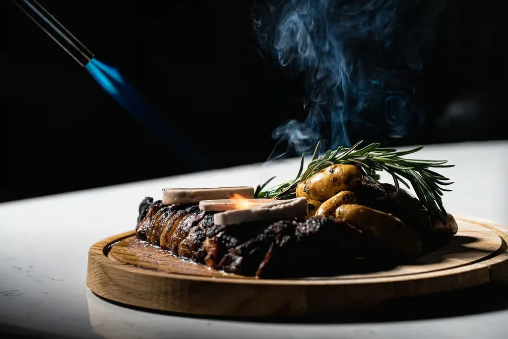 Culinary torch searing sliced meat, potatoes, and rosemary on a board.