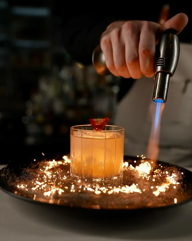 Bartender torches an amber cocktail, creating sparks on a dark tray.