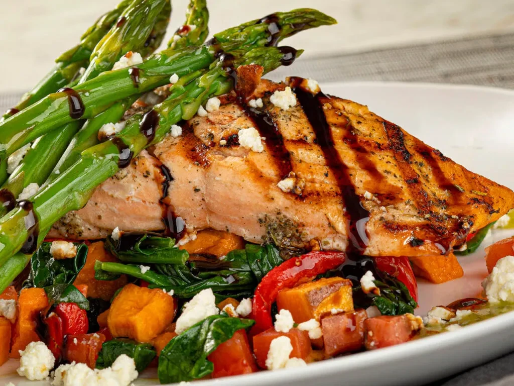 Grilled salmon with asparagus, cheese, and dark sauce on colorful vegetables.