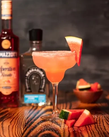 Pink watermelon margarita with salted rim, fruit garnish, blurred bottles.