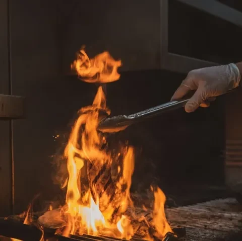 Gloved hand holds food with tongs over roaring grill fire.