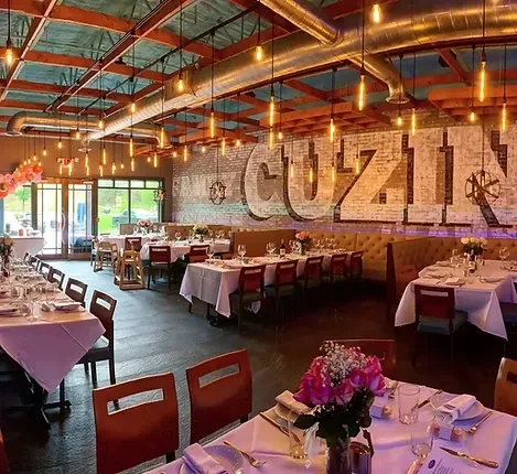 Industrial-chic restaurant with set tables, floral centerpieces, and 'CUZIN' sign.