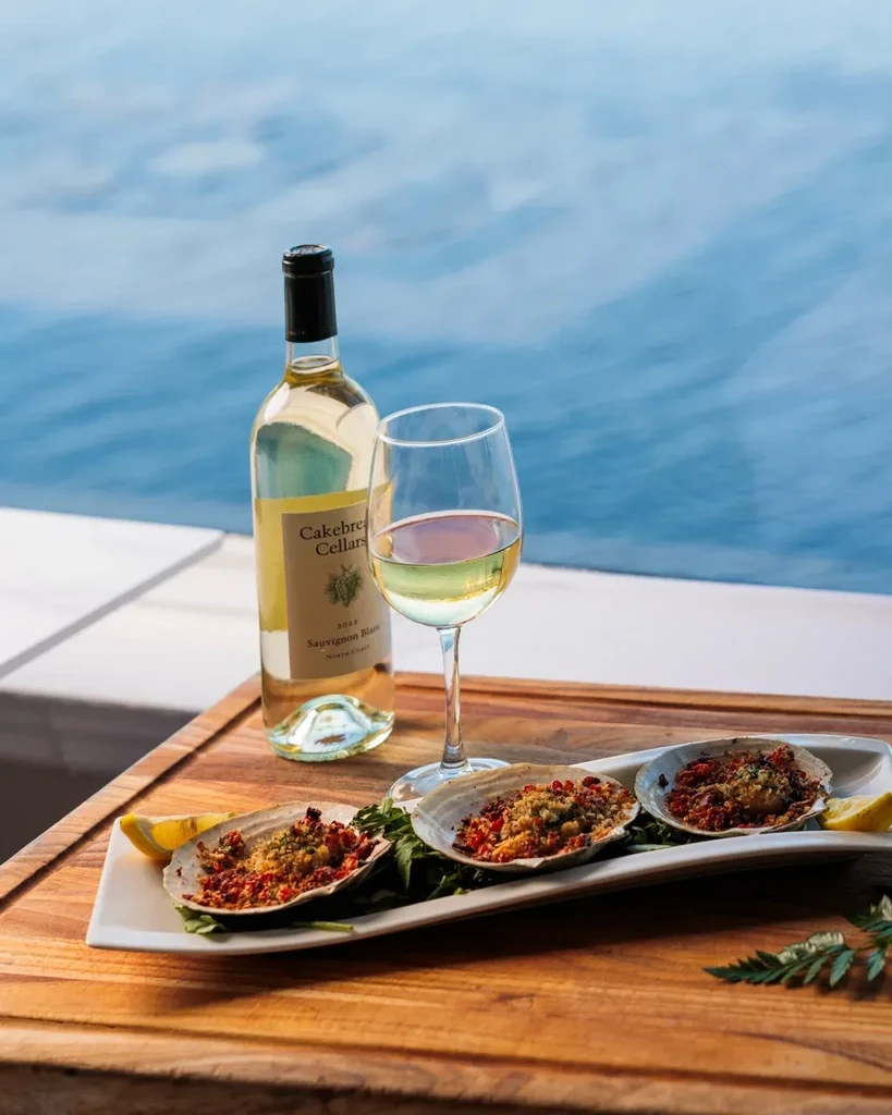 White wine bottle, baked seafood in scallop shells, serene water background.