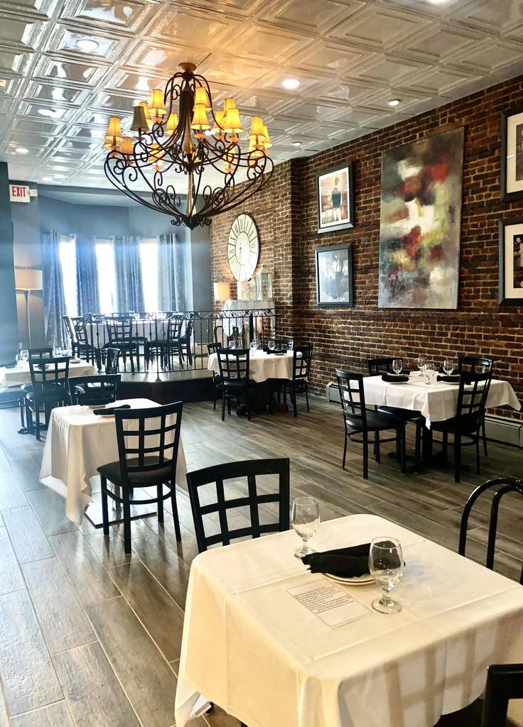 Empty dining room with set tables, white tablecloths, brick wall, chandelier.