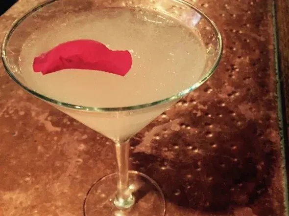 Pale cocktail with fuchsia petal in martini glass on textured surface.