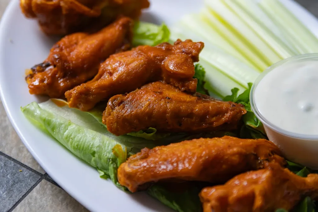 Six buffalo wings, celery, and dipping sauce on a white plate.