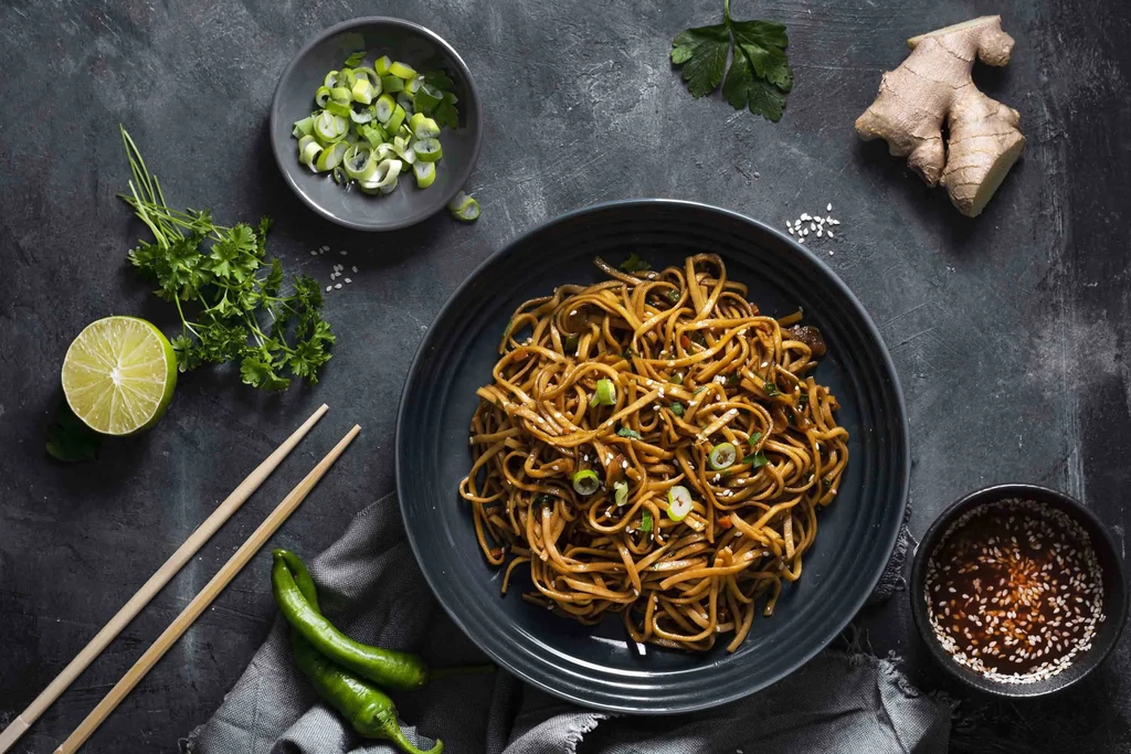 Top-down view of stir-fried noodles, garnishes, and fresh Asian ingredients.