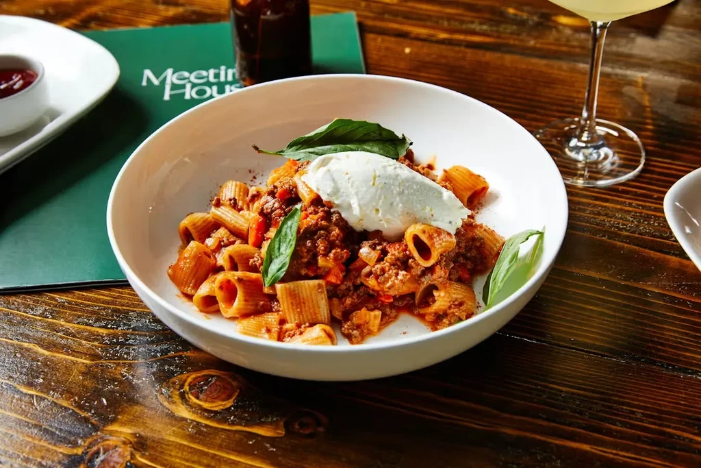 Rigatoni pasta with reddish meat sauce, creamy cheese, basil, on wood.