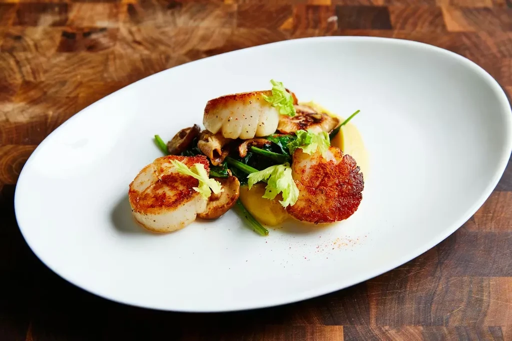 Seared scallops, yellow puree, greens, and mushrooms on a white plate.