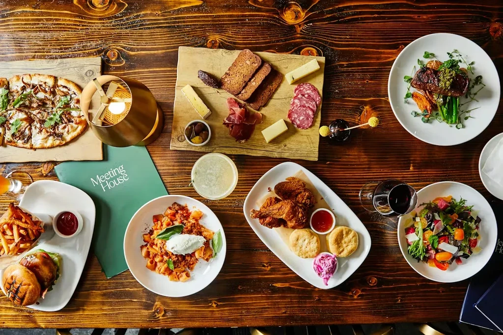 Overhead: Pizza, charcuterie, steak, pasta, fried chicken, drinks on wood table.