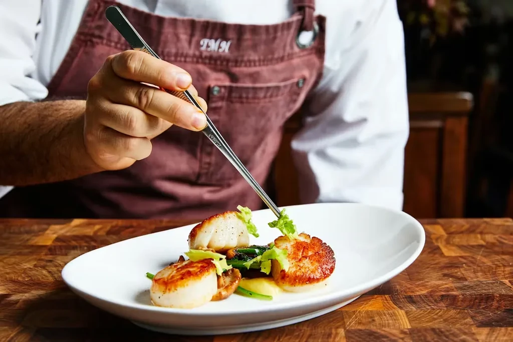 Chef's hands plating seared scallops, green vegetables, and puree.