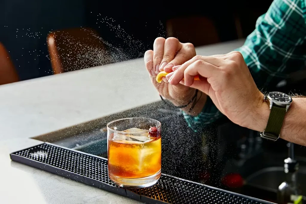 Hands squeeze citrus peel, misting an amber cocktail with a cherry.