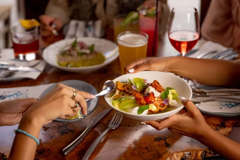 Hands share a colorful salad and drinks on a rustic wooden table.