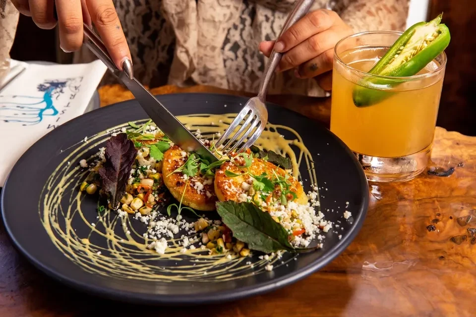 Hands cutting golden patties with cheese, herbs, beside a jalapeño cocktail.