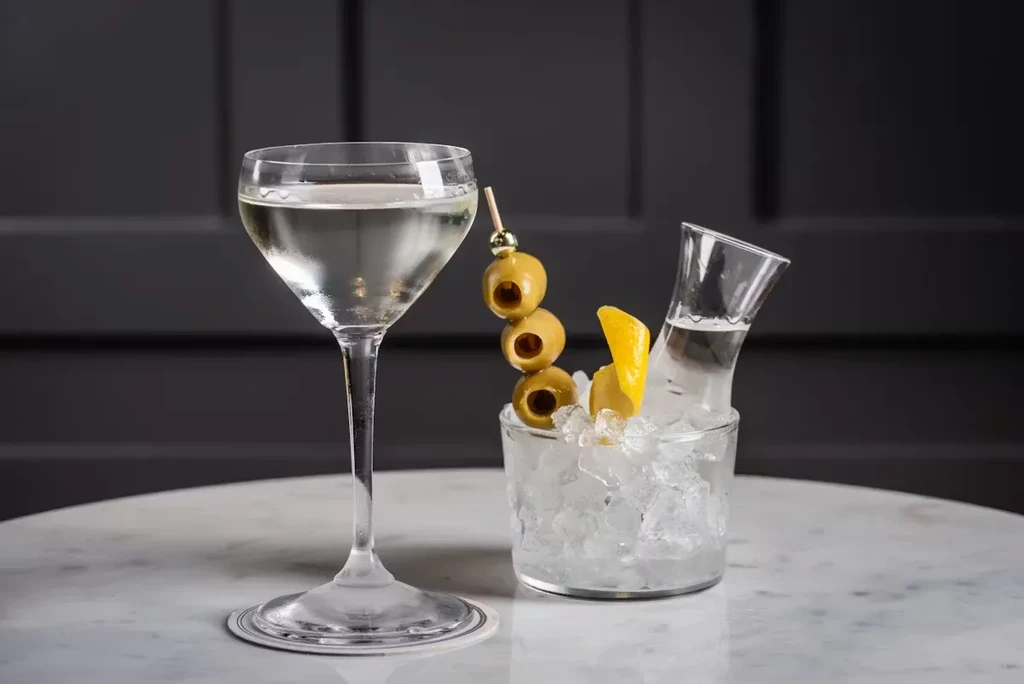 Martini, carafe in crushed ice, olives, lemon peel on marble.