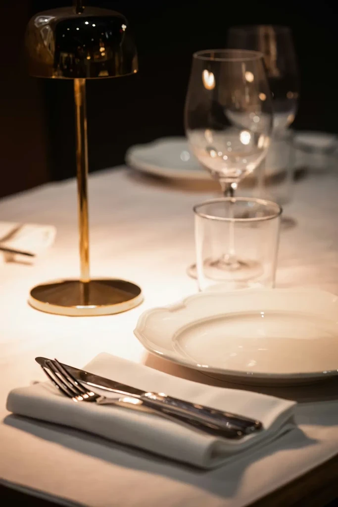Elegant restaurant table set with white plate, silver cutlery, and warm lamp.