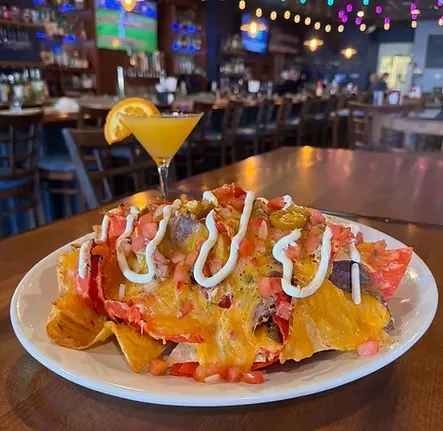 Loaded nachos, cocktail, on a wooden table in a vibrant bar.