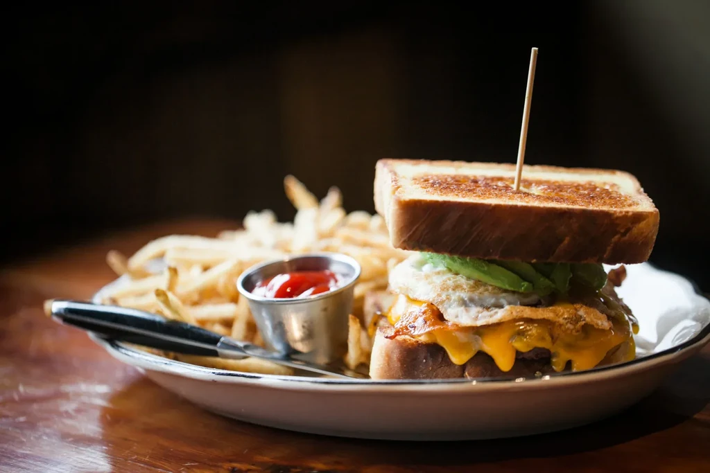 Close-up: Breakfast sandwich with egg, melted cheese, avocado, fries, ketchup.