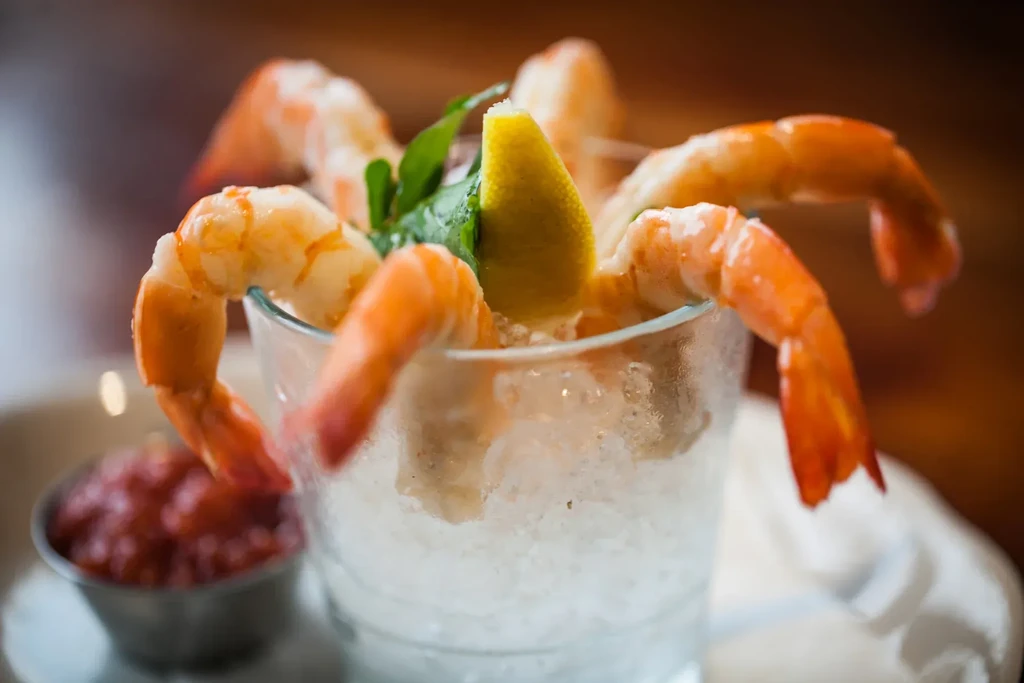 Shrimp cocktail in glass with lemon, herb, and red sauce.
